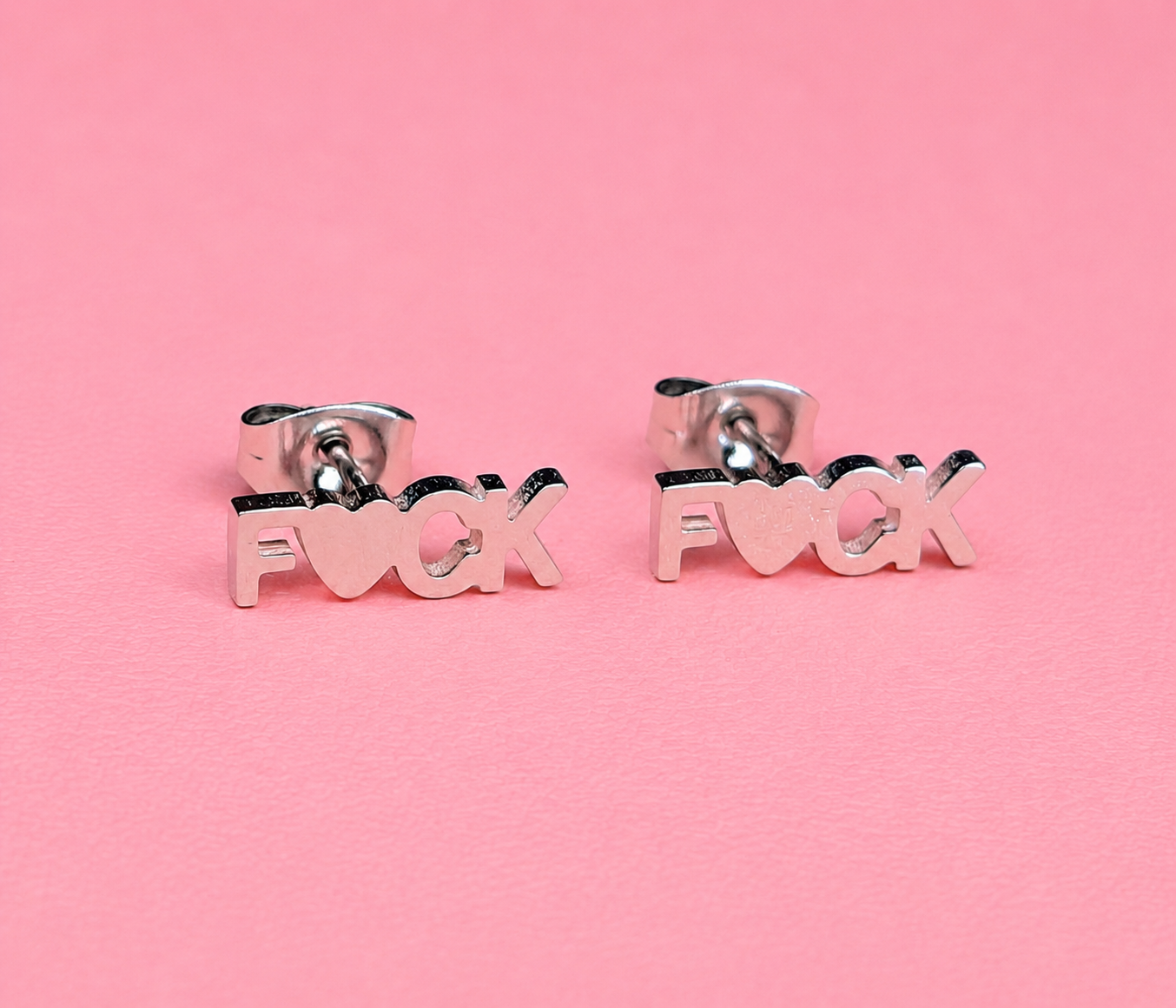 Silver 'F*ck' earrings on a pink background