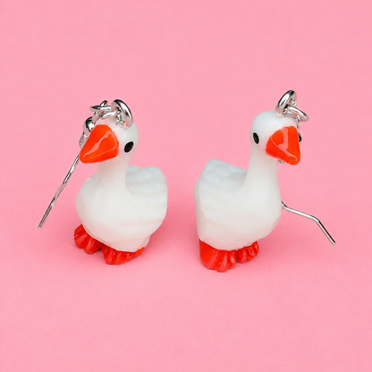 White goose-shaped earrings with orange beaks and feet on a pink background